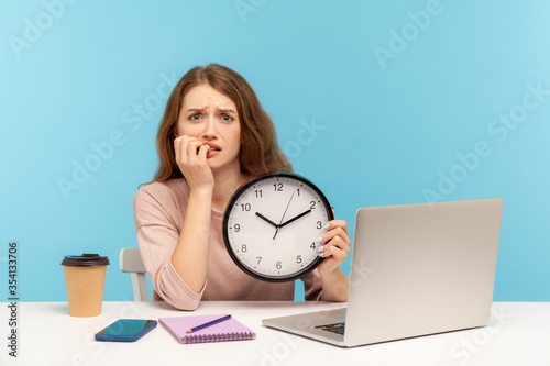 Upset anxious woman, office employee sitting at workplace, holding big clock and biting nails, working late hours, worrying about deadline, overtime job. indoor studio shot isolated on blue background