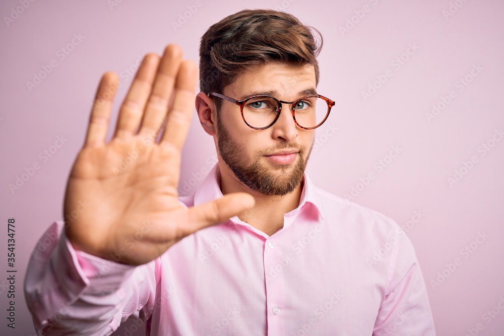 Young handsome blond man with beard and blue eyes wearing pink shirt and glasses doing stop sing with palm of the hand. Warning expression with negative and serious gesture on the face. Young handsome blond man with beard and blue eyes wearing pink shirt and glasses doing stop sing with palm of the hand. Warning expression with negative and serious gesture on the face.