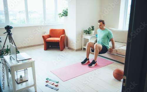 Young caucasian trainer doing fitness exercises at home having an online tutorial using camera and laptop