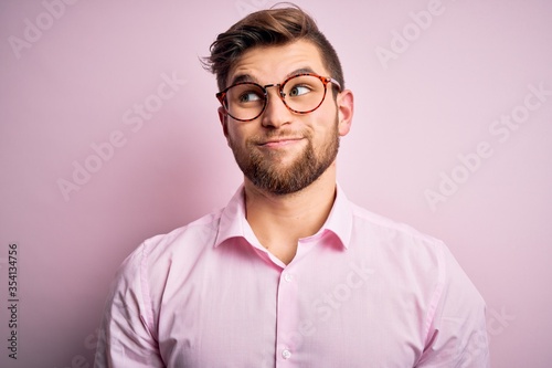 Young handsome blond man with beard and blue eyes wearing pink shirt and glasses smiling looking to the side and staring away thinking.