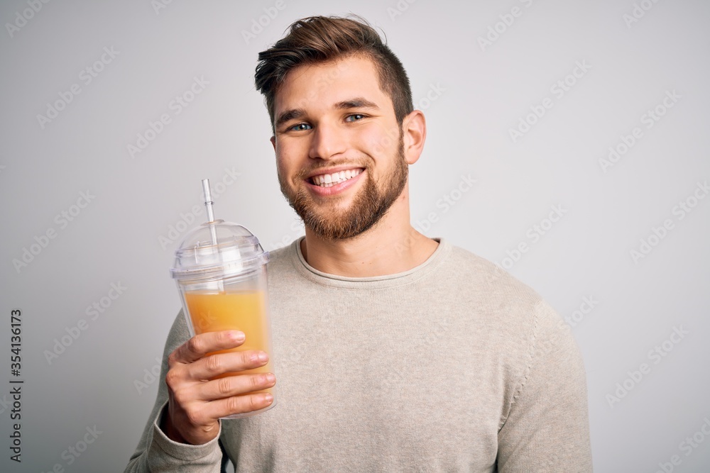 Young blond man with beard and blue eyes drinking healthy orange smoothie with a happy face standing and smiling with a confident smile showing teeth Young blond man with beard and blue eyes drinking healthy orange smoothie with a happy face standing and smiling with a confident smile showing teeth