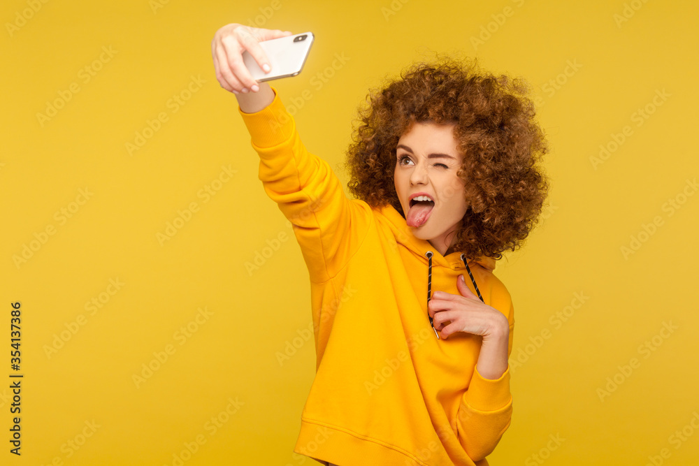 Portrait of carefree trendy curly-haired girl in urban style hoodie taking selfie and grimacing with tongue out, having fun while doing photo, vlog broadcast. studio shot isolated on yellow background Portrait of carefree trendy curly-haired girl in urban style hoodie taking selfie and grimacing with tongue out, having fun while doing photo, vlog broadcast. studio shot isolated on yellow background