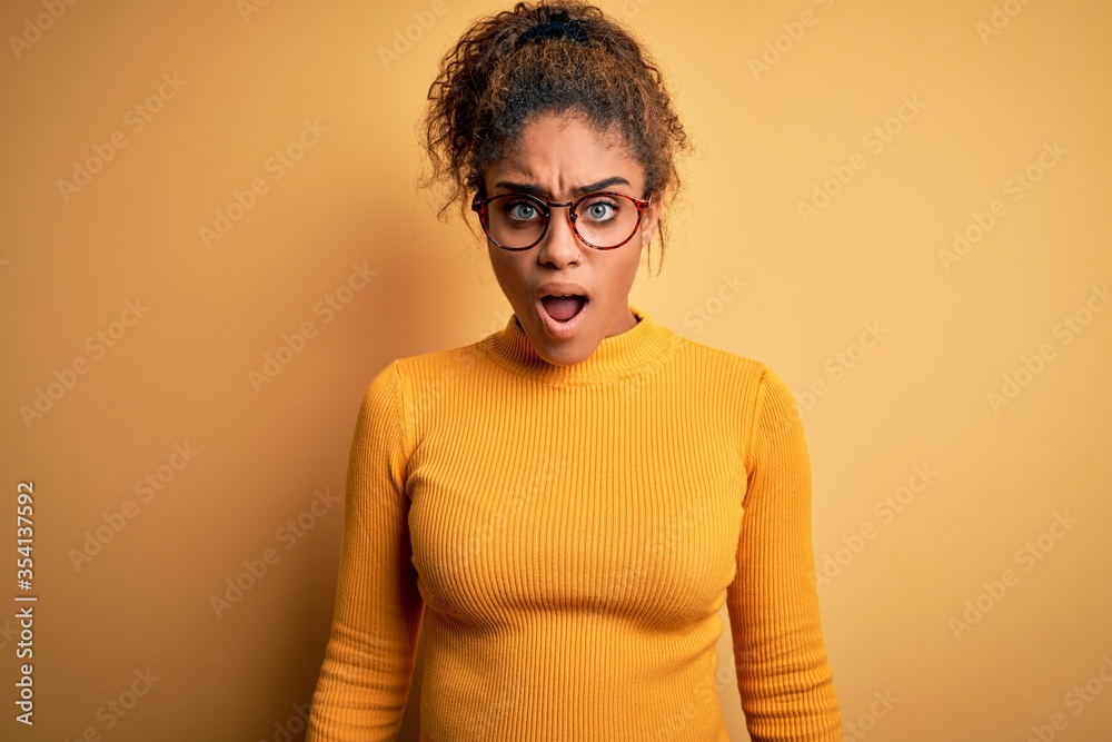 Fototapeta premium Young beautiful african american girl wearing sweater and glasses over yellow background afraid and shocked with surprise and amazed expression, fear and excited face.