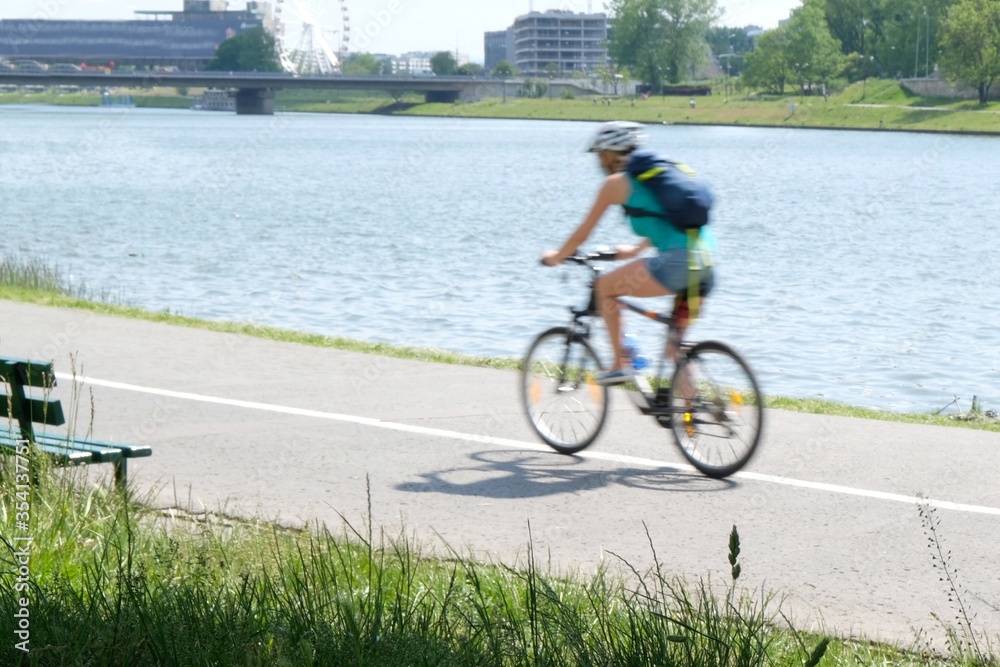 The motion blurred silhouette of a girl with a backpack rides a bicycle along a bicycle path along the Vistula river in Cracow on a warm summer day. Poland The motion blurred silhouette of a girl with a backpack rides a bicycle along a bicycle path along the Vistula river in Cracow on a warm summer day. Poland