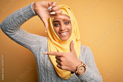 Young beautiful african american girl wearing muslim hijab over isolated yellow background smiling making frame with hands and fingers with happy face. Creativity and photography concept.