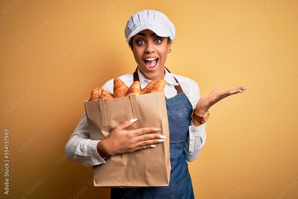 Young african american baker girl wearing apron holding bag with french croissants very happy and excited, winner expression celebrating victory screaming with big smile and raised hands Young african american baker girl wearing apron holding bag with french croissants very happy and excited, winner expression celebrating victory screaming with big smile and raised hands