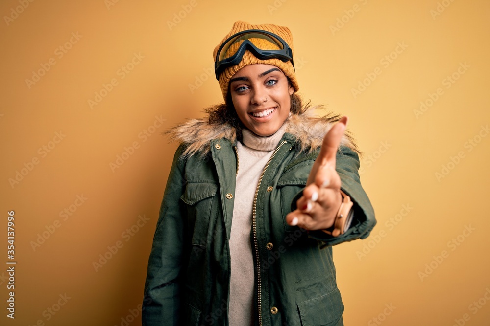 Fototapeta premium Young african american afro skier girl wearing snow sportswear and ski goggles smiling cheerful offering palm hand giving assistance and acceptance.