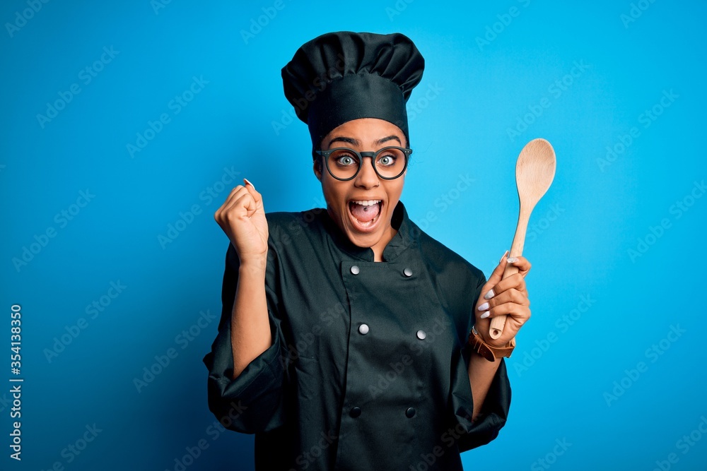 Fototapeta premium Young african american chef girl wearing cooker uniform and hat holding wooden spoon tool screaming proud and celebrating victory and success very excited, cheering emotion
