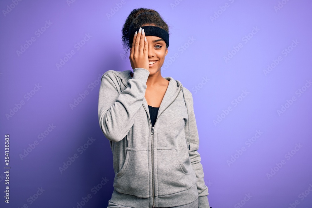 Young african american sportswoman doing sport wearing sportswear over purple background covering one eye with hand, confident smile on face and surprise emotion.