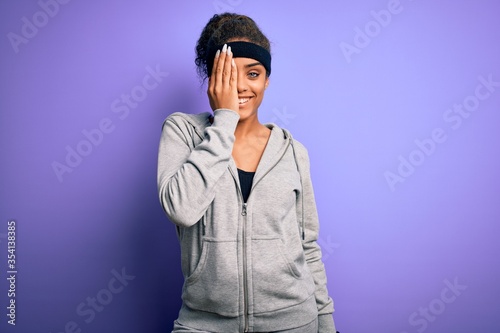 Young african american sportswoman doing sport wearing sportswear over purple background covering one eye with hand  confident smile on face and surprise emotion.