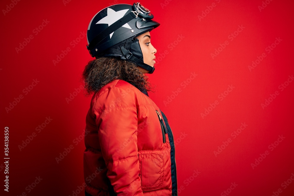 Young african american motorcyclist girl wearing moto helmet and glasses over red background looking to side, relax profile pose with natural face with confident smile. Young african american motorcyclist girl wearing moto helmet and glasses over red background looking to side, relax profile pose with natural face with confident smile.