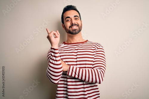 Young handsome man with beard wearing casual striped t-shirt standing over white background with a big smile on face  pointing with hand and finger to the side looking at the camera.