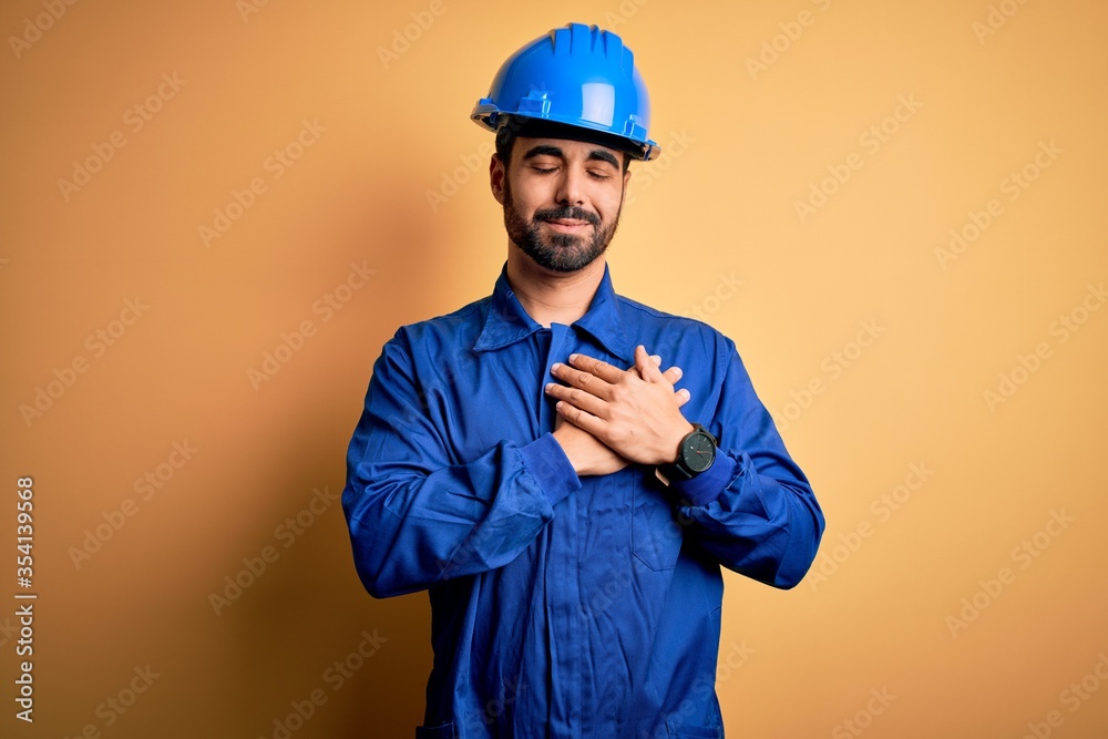 Mechanic man with beard wearing blue uniform and safety helmet over yellow background smiling with hands on chest with closed eyes and grateful gesture on face. Health concept.
