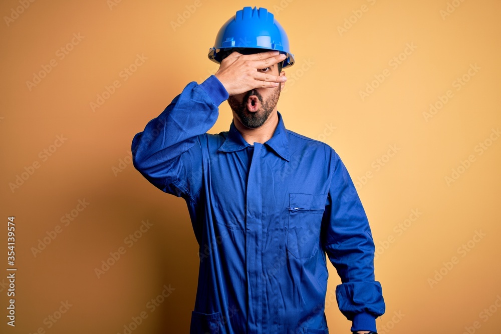 Mechanic man with beard wearing blue uniform and safety helmet over yellow background peeking in shock covering face and eyes with hand, looking through fingers with embarrassed expression.