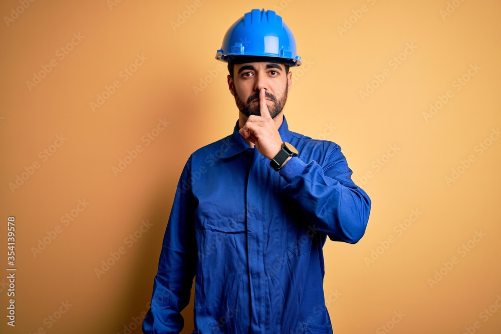 Mechanic man with beard wearing blue uniform and safety helmet over yellow background asking to be quiet with finger on lips. Silence and secret concept.