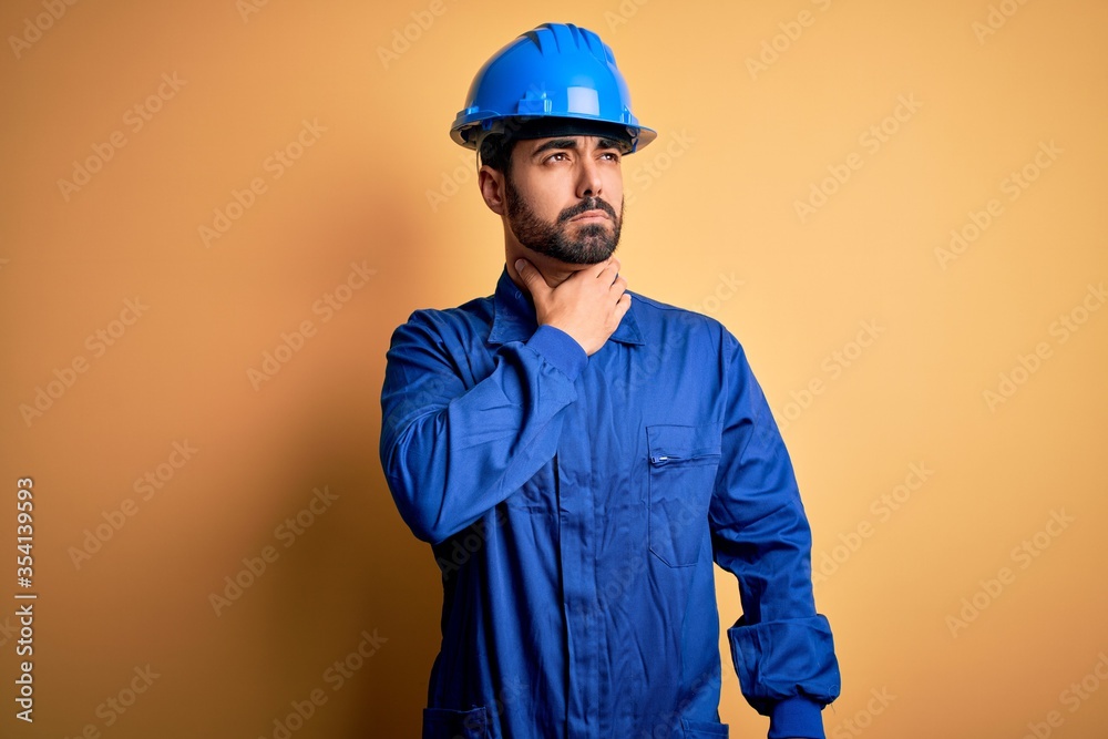 Mechanic man with beard wearing blue uniform and safety helmet over yellow background Touching painful neck, sore throat for flu, clod and infection