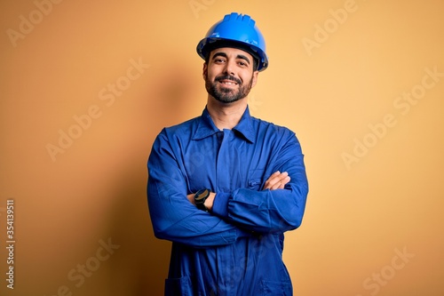 Mechanic man with beard wearing blue uniform and safety helmet over yellow background happy face smiling with crossed arms looking at the camera. Positive person.
