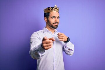 Young handsome man with beard wearing golden crown of king over purple background pointing fingers to camera with happy and funny face. Good energy and vibes.