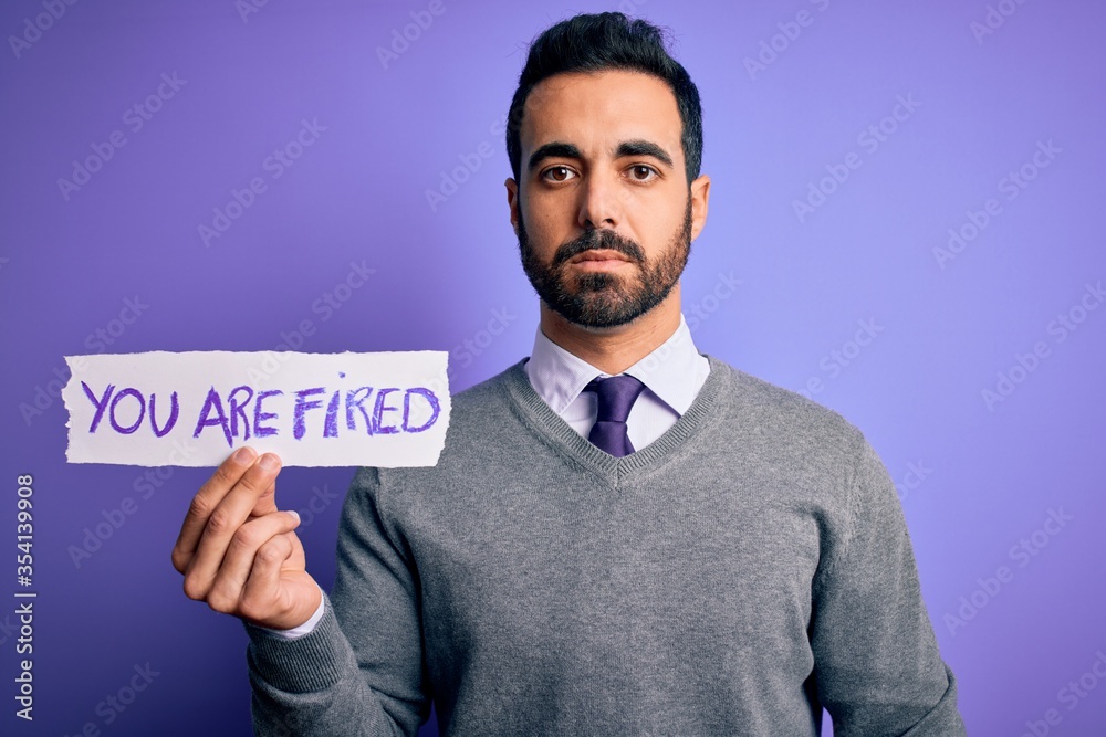 Young handsome businessman with beard holding paper with you are fired message with a confident expression on smart face thinking serious Young handsome businessman with beard holding paper with you are fired message with a confident expression on smart face thinking serious