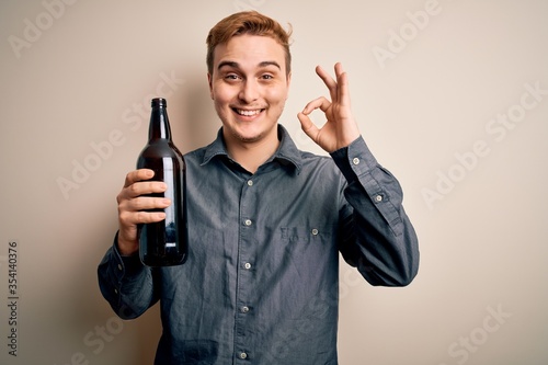 Young handsome redhead man drinking bottle of beer over isolated white background doing ok sign with fingers, smiling friendly gesturing excellent symbol