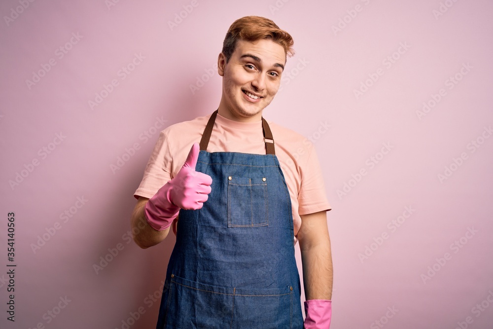 Young handsome redhead cleaner man doing housework wearing apron and gloves doing happy thumbs up gesture with hand. Approving expression looking at the camera showing success. Young handsome redhead cleaner man doing housework wearing apron and gloves doing happy thumbs up gesture with hand. Approving expression looking at the camera showing success.