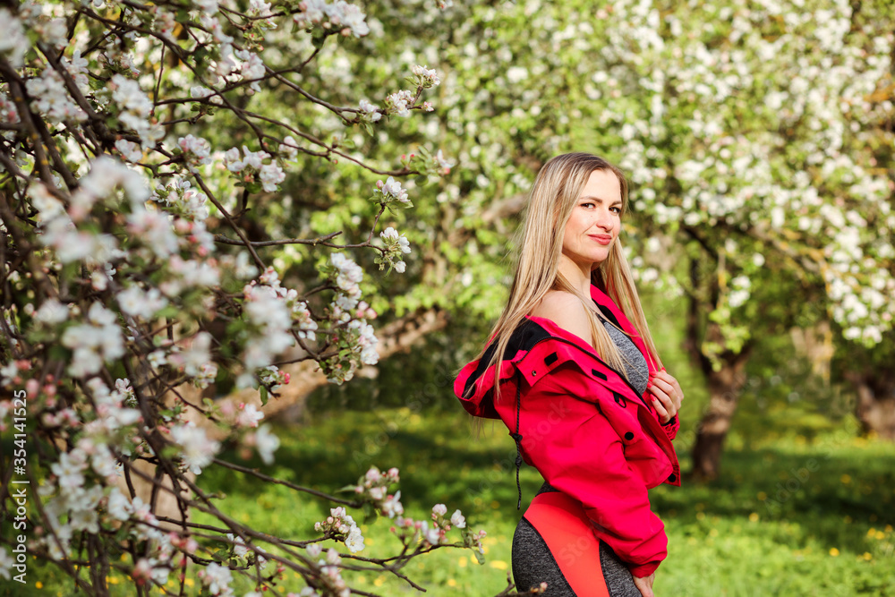 Portrait of the caucasian woman wearing pink bomber jacket and tights outdoors in the blooming park in spring. Beautiful blonde woman spending her leisure outside in the blooming garden.