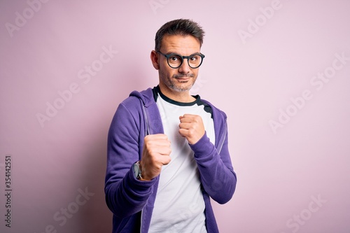 Young handsome man wearing purple sweatshirt and glasses standing over pink background Ready to fight with fist defense gesture, angry and upset face, afraid of problem
