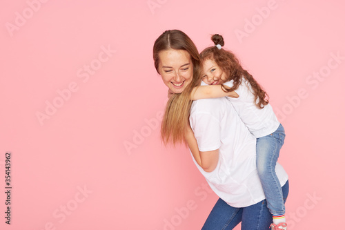 Mom and daughter in white t-shirts and jeans play and hug on a pink background. Caring for loved ones. Happy motherhood