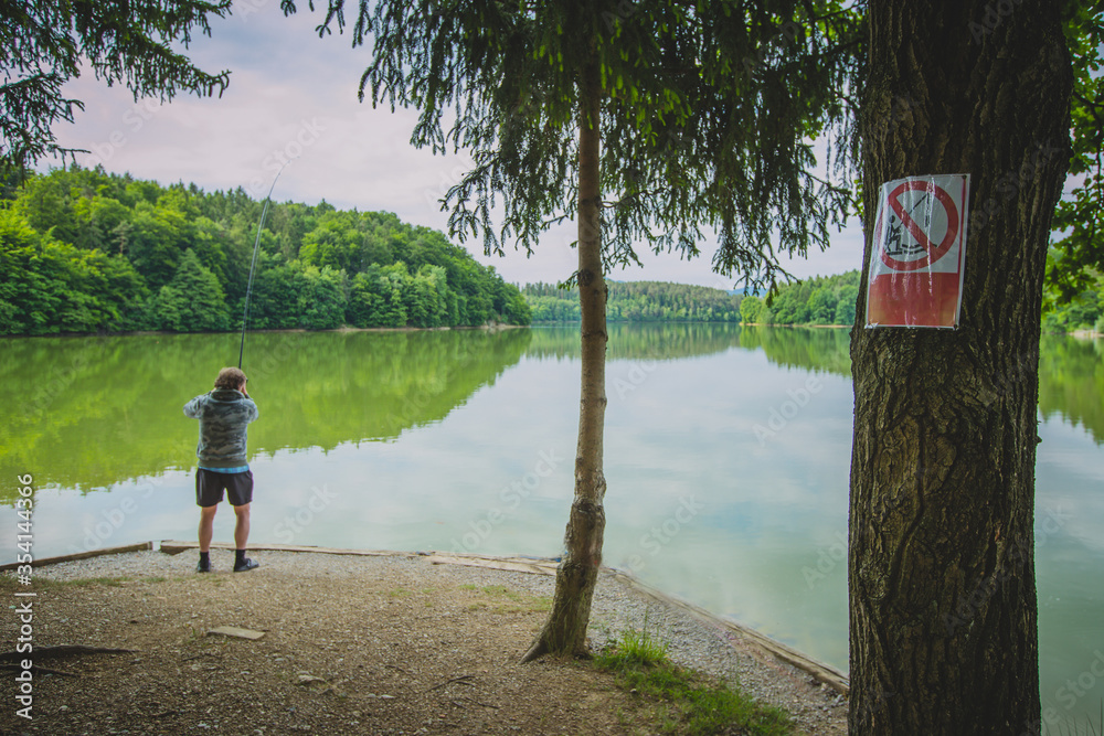 Man is fishing on a place at a lake where it is clearly stated that you shouldn't as the sign on the tree is prohibiting it.