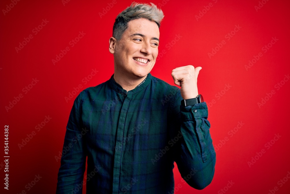 Young handsome modern man wearing elegant green shirt over red isolated background smiling with happy face looking and pointing to the side with thumb up.