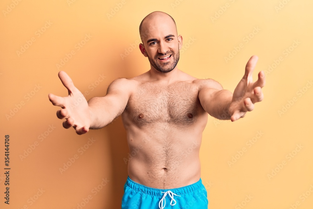 Young handsome bald man wearing swimwear shirtless looking at the camera smiling with open arms for hug. cheerful expression embracing happiness.