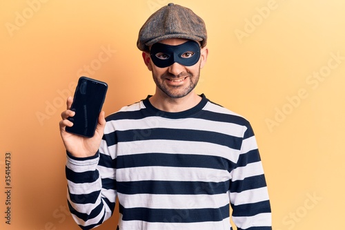 Young handsome bald man wearing burglar mask holding smartphone looking positive and happy standing and smiling with a confident smile showing teeth © Krakenimages.com