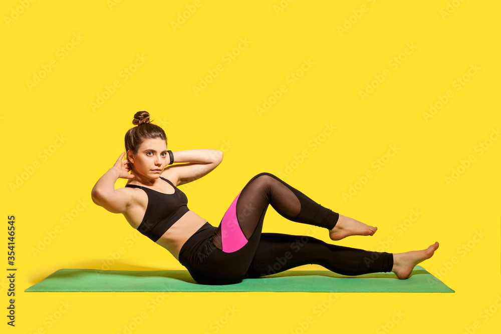 Obraz premium Fitness young woman with hair bun in tight sportswear training on gym mat, doing sit ups crunch, exercising abdominal muscles, lower-body workouts, warming up. indoor studio shot, isolated on yellow