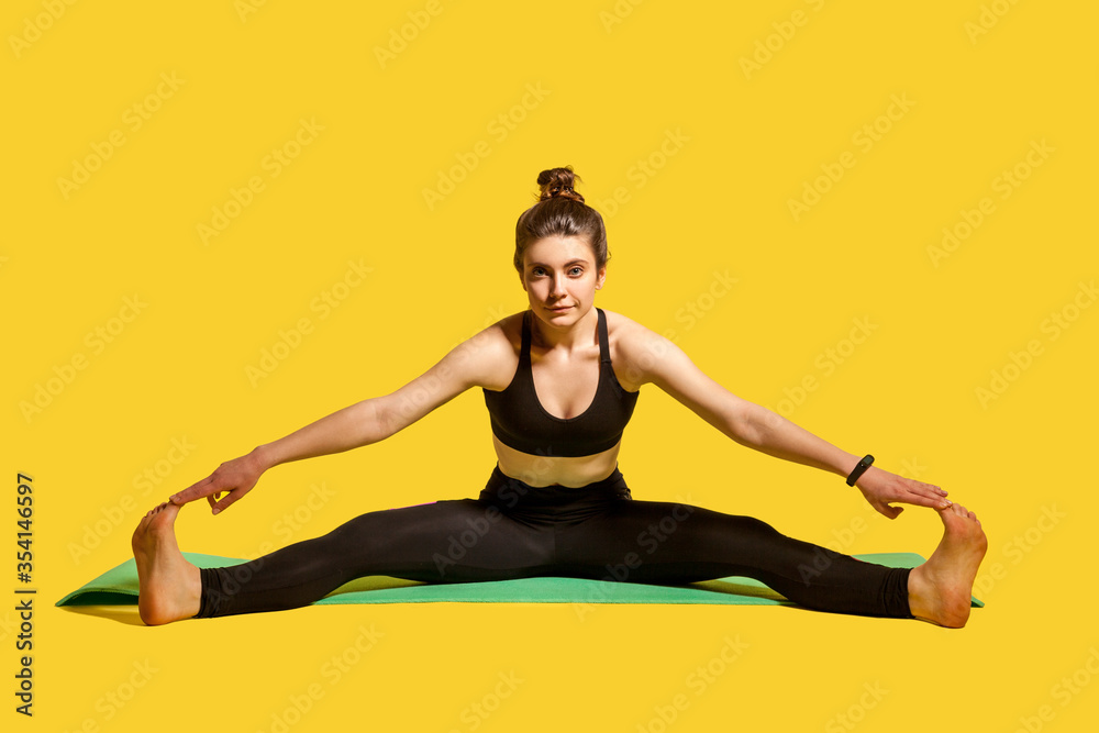 Gymnast woman with hair bun in tight sportswear sitting on mat with spread legs, touching toes stretching muscles, doing sports flexibility exercises. indoor studio shot isolated on yellow background Gymnast woman with hair bun in tight sportswear sitting on mat with spread legs, touching toes stretching muscles, doing sports flexibility exercises. indoor studio shot isolated on yellow background