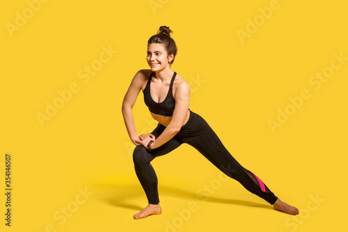 Happy athletic girl with hair bun in tight sportswear doing lower body sport exercise  stretching legs  warming up training muscles for flexibility. indoor full length studio shot  isolated on yellow