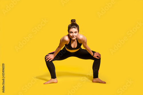 Motivated assertive gymnast girl with hair bun in tight sportswear squatting and spreading legs to stretch muscles  warming up before twine exercise  training flexibility. full length studio shot