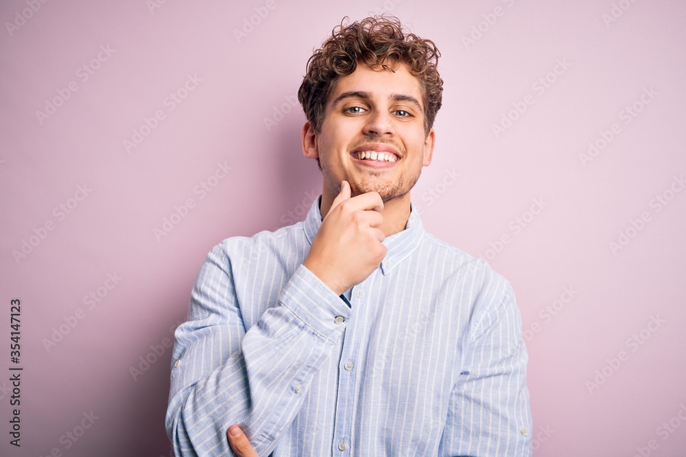 Young blond handsome man with curly hair wearing striped shirt over white background looking confident at the camera smiling with crossed arms and hand raised on chin. Thinking positive. Young blond handsome man with curly hair wearing striped shirt over white background looking confident at the camera smiling with crossed arms and hand raised on chin. Thinking positive.
