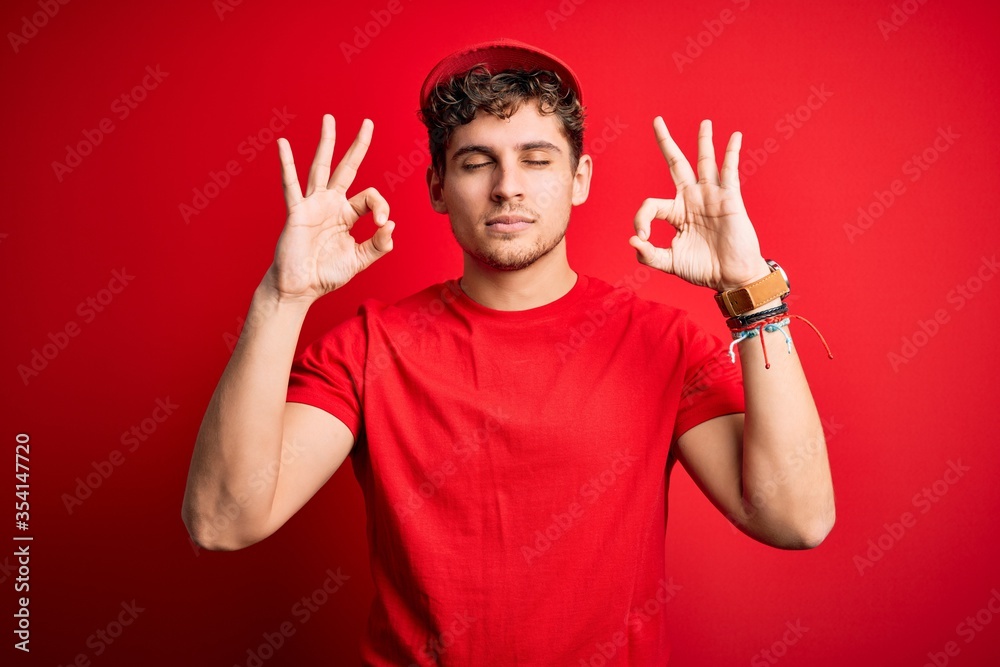 Young blond delivery man with curly hair wearing cap standing over isolated red background relax and smiling with eyes closed doing meditation gesture with fingers. Yoga concept. Young blond delivery man with curly hair wearing cap standing over isolated red background relax and smiling with eyes closed doing meditation gesture with fingers. Yoga concept.