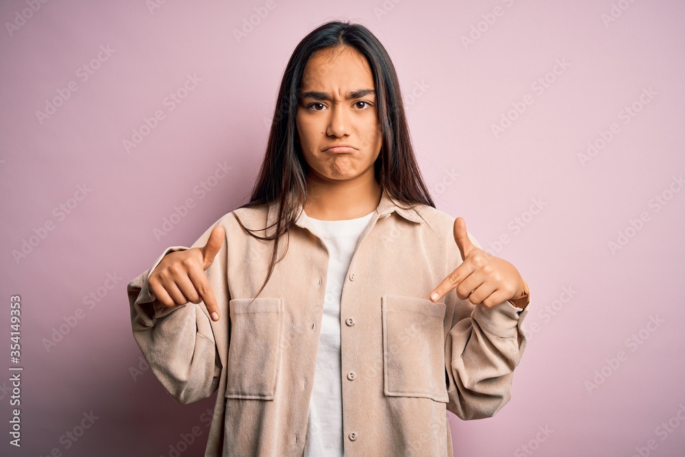 Young beautiful asian woman wearing casual shirt standing over pink background Pointing down looking sad and upset, indicating direction with fingers, unhappy and depressed. Young beautiful asian woman wearing casual shirt standing over pink background Pointing down looking sad and upset, indicating direction with fingers, unhappy and depressed.
