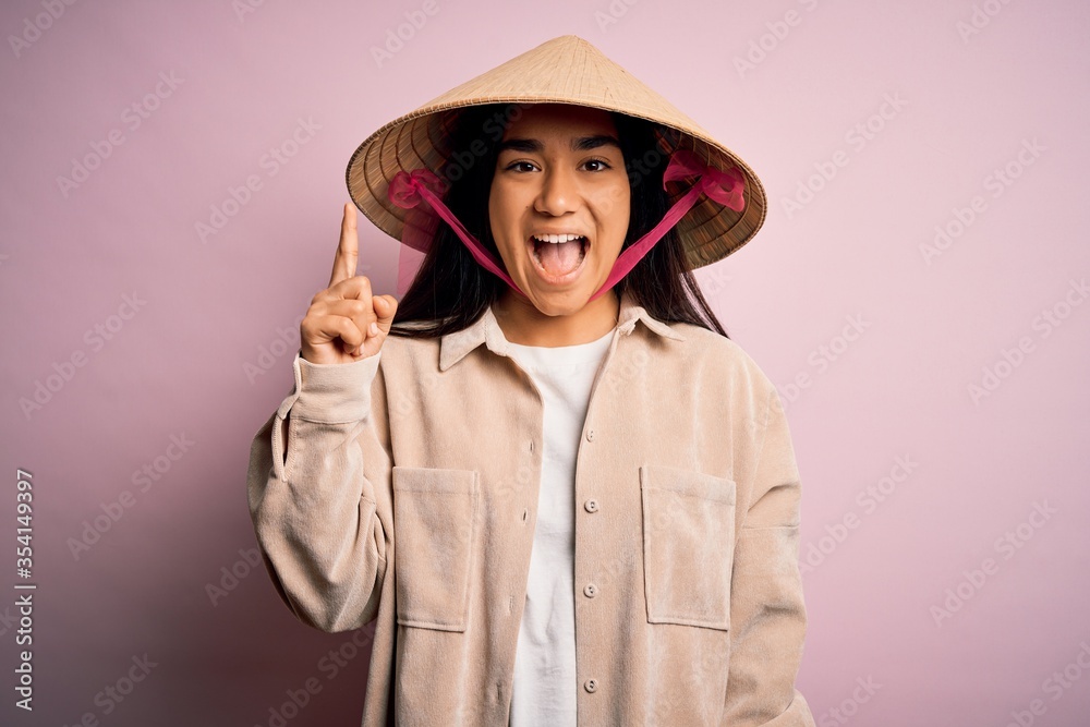 Young beautiful woman wearing traditional conical asian hat over isolated pink background pointing finger up with successful idea. Exited and happy. Number one. Young beautiful woman wearing traditional conical asian hat over isolated pink background pointing finger up with successful idea. Exited and happy. Number one.