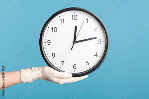 Profile side view closeup of human hand in white surgical gloves holding analog clock. indoor, studio shot, isolated on blue background.