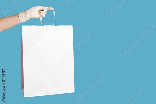 Profile side view closeup of human hand in white surgical gloves holding and showing shopping bags. indoor, studio shot, isolated on blue background.
