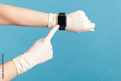 Profile side view closeup of human hand in white surgical gloves holding and showing wirst smart watch and pointing at empty screen. indoor, studio shot, isolated on blue background. photo