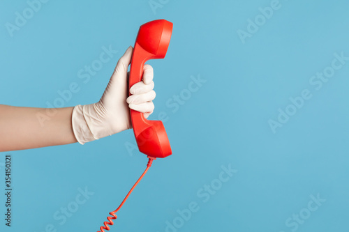 Profile side view closeup of human hand in white surgical gloves holding and showing red cale telephone handset receiver. indoor, studio shot, isolated on blue background.