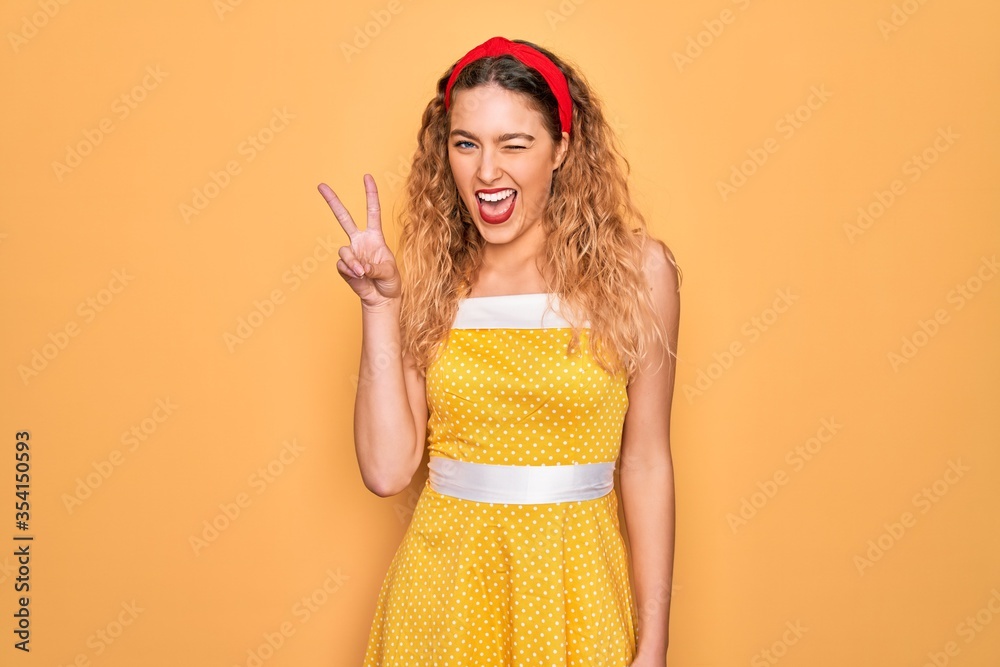 Beautiful blonde pin-up woman with blue eyes wearing diadem standing over yellow background smiling with happy face winking at the camera doing victory sign with fingers. Number two. Beautiful blonde pin-up woman with blue eyes wearing diadem standing over yellow background smiling with happy face winking at the camera doing victory sign with fingers. Number two.