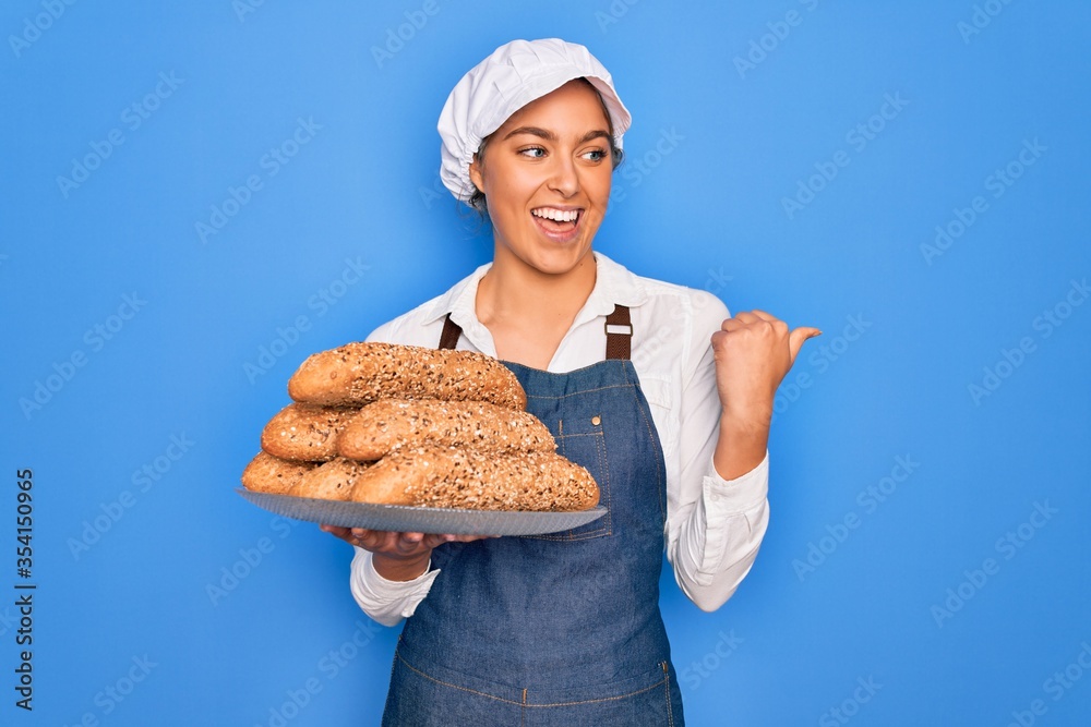 Young beautiful blonde baker woman with blue eyes holding tray with wholemeal bread cereal pointing and showing with thumb up to the side with happy face smiling Young beautiful blonde baker woman with blue eyes holding tray with wholemeal bread cereal pointing and showing with thumb up to the side with happy face smiling
