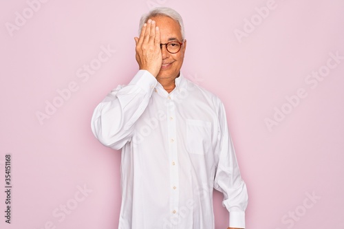 Middle age senior grey-haired man wearing glasses and business shirt over pink background covering one eye with hand, confident smile on face and surprise emotion.