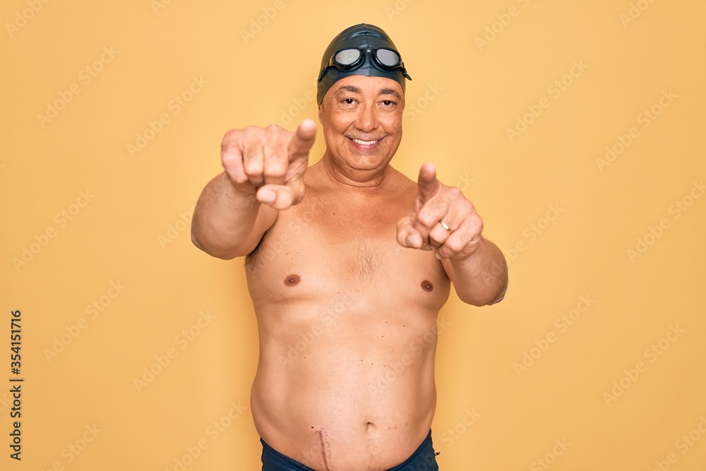Middle age senior grey-haired swimmer man wearing swimsuit, cap and goggles pointing to you and the camera with fingers, smiling positive and cheerful