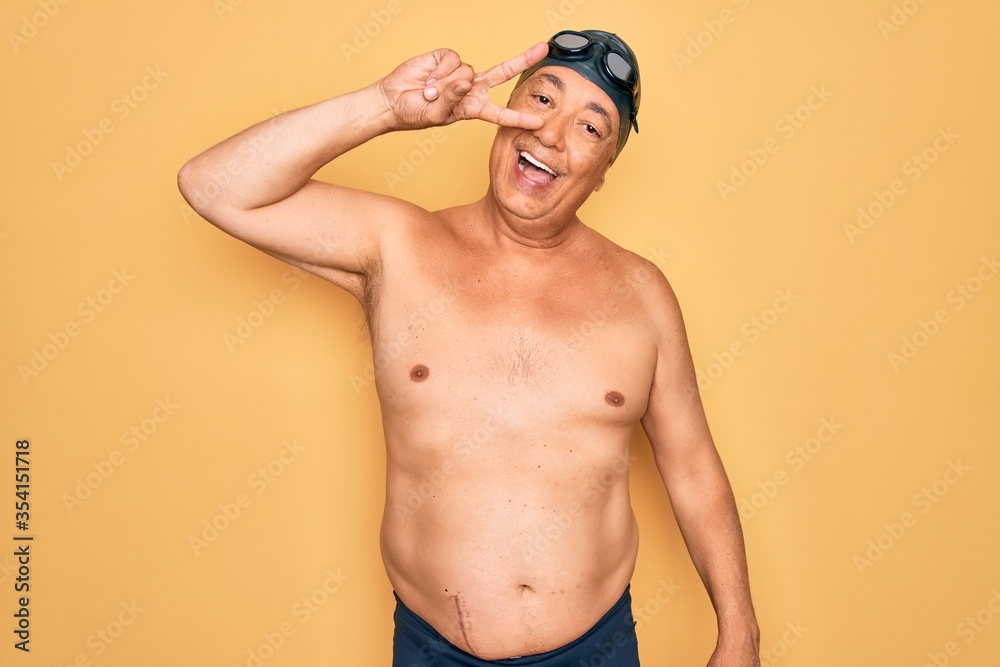 Middle age senior grey-haired swimmer man wearing swimsuit, cap and goggles Doing peace symbol with fingers over face, smiling cheerful showing victory