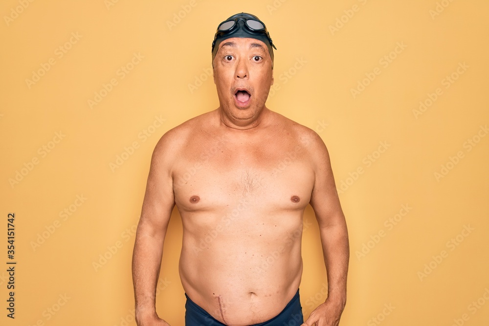 Middle age senior grey-haired swimmer man wearing swimsuit, cap and goggles afraid and shocked with surprise and amazed expression, fear and excited face.